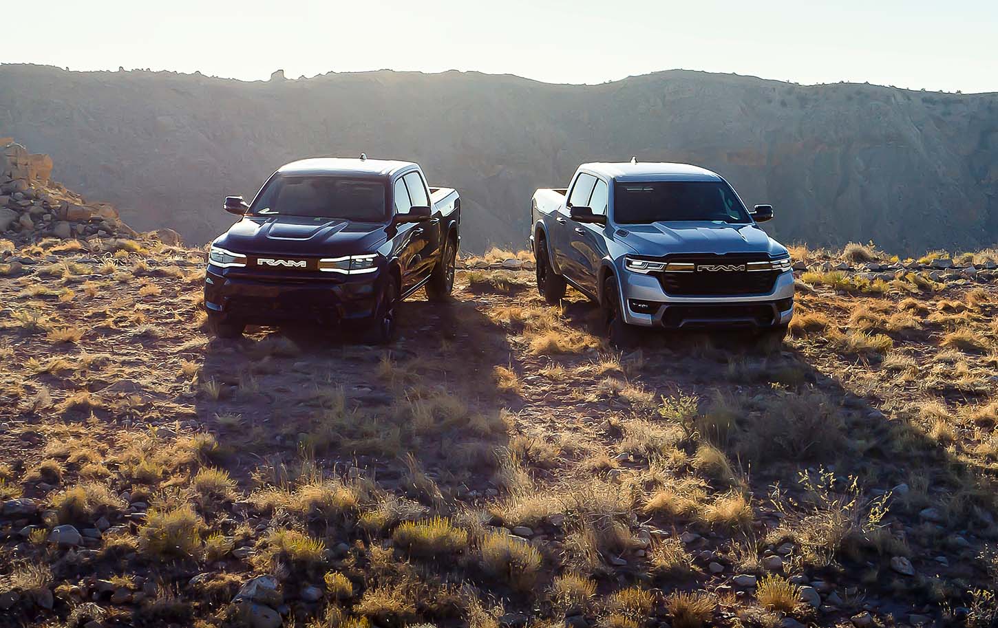 A gray 2025 Ram 1500 Rev Tungsten Crew Cab and a silver 2025 Ram 1500 Ramcharger Limited Crew Cab parked on a clearing in the mountains.