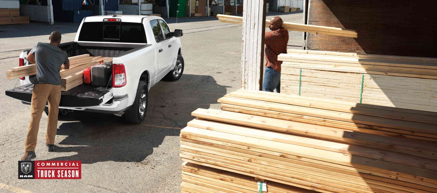 A rear angle of a white 2024 Ram 1500 Tradesman Crew Cab parked at a lumber yard with its tailgate open and a man loading lumber into the truck bed.