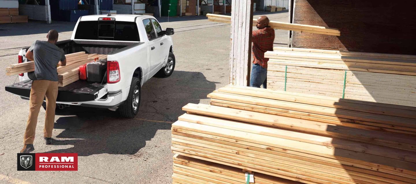 Ángulo trasero de una Ram 1500 Tradesman Crew Cab 2024 blanca estacionada en un depósito de madera con la puerta trasera abierta y un hombre cargando madera en la plataforma de la camioneta mientras otro hombre trae más madera. Ram Professional.