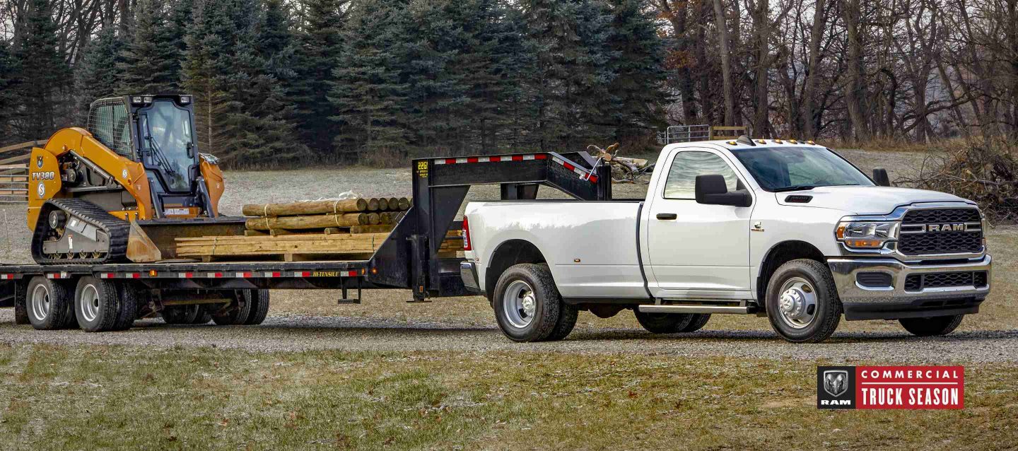 A white 2024 Ram 3500 Tradesman 4x4 Regular Cab towing a fifth wheel flatbed trailer loaded with an excavator and lumber.