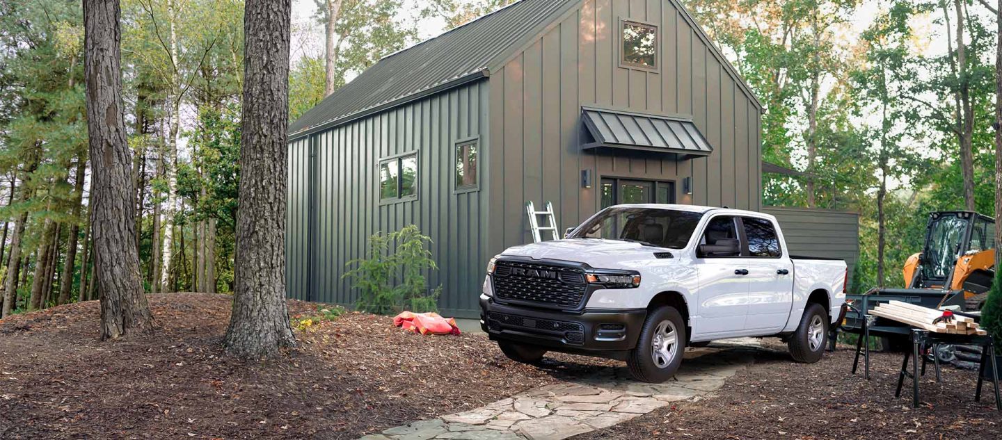 Una Ram 1500 Tradesman 4 x 4 2025 blanca estacionada al lado de una cabaña en el bosque.