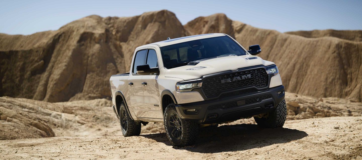 Vista en ángulo delantera del lado del pasajero de una Ram 1500 Rebel X Crew Cab 2025 blanca circulando por una colina en el desierto, fuera de la carretera.