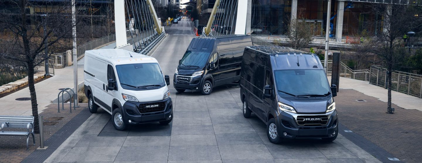 Tres modelos Ram ProMaster 2025 aparcados al otro lado de un puente en el centro de una gran ciudad, de izquierda a derecha: una Ram ProMaster 1500 Tradesman Cargo Van Standard Roof blanca, una Ram ProMaster 3500 SLT Plus Cargo Van Super High Roof negra y una Ram ProMaster 3500 SLT Cargo Van High Roof negra.