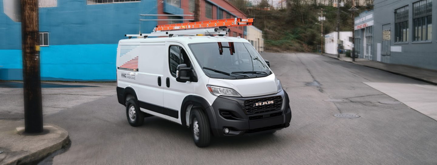 Una Ram ProMaster 1500 Tradesman Cargo Van Standard Roof blanca de 2025 con el logotipo de una empresa de climatización en el lateral y una escalera en la baca girando a la derecha por una zona industrial.