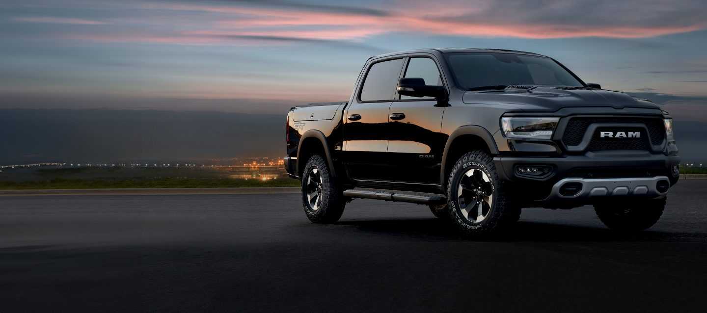 A front angled passenger-side profile of a blue 2024 Ram 1500 Limited Crew Cab parked in the driveway of a large home. Power Days Sales Event.